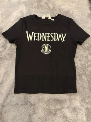 Svart Wednesday t-shirt från H&M - Cool svart t-shirt från H&M med trycket 'WEDNESDAY' och ett emblem med texten 'Unitas est invicta' på bröstet. T-shirten har korta ärmar, rund hals och är gjord i mjuk bomull. Perfekt för dig som gillar Wednesday eller vill ha en statement-tee.