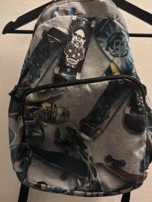 Ryggsäck med skateboardmönster - Cool ryggsäck med grå bas och tryck av olika skateboards i blå, svart och gröna toner. Väskan har ett stort huvudfack och en praktisk ytterficka med dragkedja. 