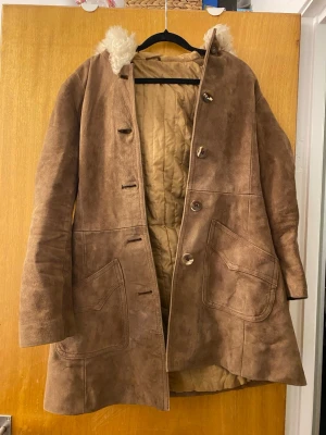 Brun mocka kappa, vintage - Säljer en brun kappa i mocka från Aùlùna, tillverkad i Danmark. Jackan har stor huva med fluffig päls, stora fickor framtill, knappar och bälte i midjan. Insidan är quiltad för extra värme och den har en riktig vintage-vibe.