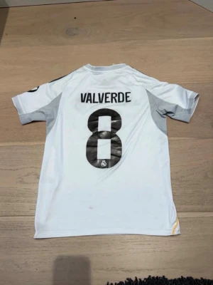 Real Madrid  matchtröja - Vit Real Madrid matchtröja från Adidas med Valverde och nummer 8 på ryggen. Tröjan har Champions League-märke på ärmen, bruna ränder på axlarna och gula detaljer längs sidorna. Materialet är polyester och modellen är kortärmad.