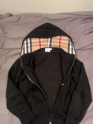 Burberry zip up black  - Burberry hoodie zip Svart med klassiskt rutigt foder  Stilren svart Burberry hoodie med dragkedja. Insidan av huvan är fodrad med Burberrys ikoniska beige rutmönster, vilket ger en exklusiv och tidlös detalj. Mjuk och bekväm kvalitet som passar perfekt för både vardag och mer uppklädda tillfällen.  Jackan har en klassisk passform, huva med dragsko och genomgående dragkedja framtill. Ett elegant plagg från Burberry som kombinerar komfort med lyxig design.