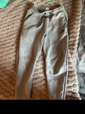 Grå mjukisbyxor från Nike XS - Säljer ett par grå mjukisbyxor från Nike i storlek XS. Byxorna har dragsko i midjan, ribbade muddar vid bensluten och två fickor. Tillverkade i mjukt bomullsmaterial och har Nike-logga broderad på vänster sida.
