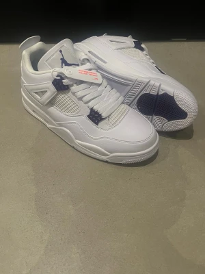 Nike Air Jordan 4 vit/lila sneakers - Nike Air Jordan 4 sneakers i vitt skinn med lila detaljer och klassisk Jumpman-logga på plös och häl. Skorna har meshpaneler, snörning och en chunky sula för extra streetkänsla. Perfekta för dig som vill ha en ikonisk och stilren sneaker.