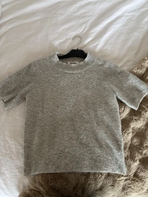 Grå stickad tröja från Missäy - Mysig grå stickad tröja från Missäy med korta ärmar och ribbade detaljer vid hals, ärmslut och nederkant. Perfekt till lager på lager och enkel att matcha med allt. Klassisk och stilren modell.