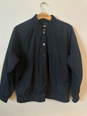 Svart bomberjacka med knappar - Säljer en svart bomberjacka från Weekday med klassisk ribbad krage, muddar och knappstängning framtill. Jackan har en clean look utan synliga loggor och är tillverkad i ett lätt, syntetmaterial. Perfekt för dig som gillar minimalistisk stil.