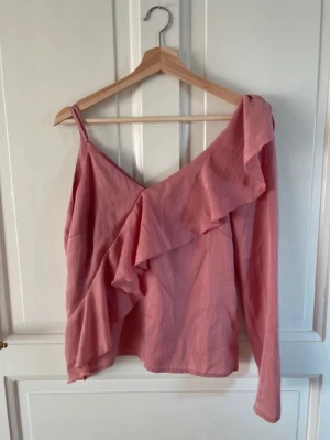 Rosa omlottopp med volang - Säljer en rosa omlottopp med en axelbandslös sida och en lång ärm. Toppen har en snygg volangdetalj som går diagonalt över framsidan och ger en cool asymmetrisk look.