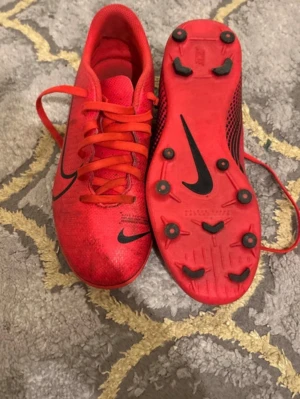 Röda Nike fotbollsskor med dobbar - Säljer ett par röda Nike fotbollsskor med svarta detaljer och klassisk swoosh-logga. Skorna har snörning och rund tå samt slitstarka dobbar för bra grepp på planen. Ovandelen är i syntetmaterial som ger en lätt känsla och bra bollkontroll.