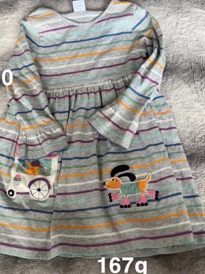 Randig barnklänning med hundmotiv - Superfin barnklänning med färgglada ränder. Klänningen har trekvarts ärm och är dekorerad med ett gulligt hundmotiv och en ficka med glassbil. perfekt för lek och bus.🌸 Små fläckar. Storlek 110