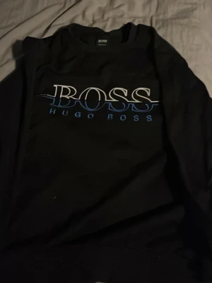 Svart Hugo Boss sweatshirt slim fit - Säljer en svart sweatshirt från Hugo Boss med stor BOSS-logga i vitt och blått framtill. Tröjan har rund halsringning och är i slim fit-modell. Perfekt för dig som gillar stilrena och klassiska plagg med en modern touch.