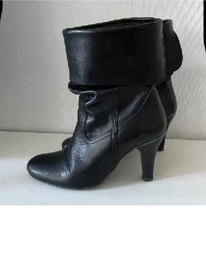 Svarta stövletter i skinn med klack - Säljer ett par svarta stövletter i skinn med vikbar kant och smal klack. Skorna har en elegant, spetsig tå och en klassisk design som passar till många olika outfits. Perfekta för dig som gillar stilrena boots med lite höjd.