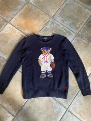 Mörkblå stickad tröja Polo Ralph Lauren - Mörkblå stickad tröja från Polo Ralph Lauren med ikonisk björn i basebolloutfit broderad på bröstet. RL-broderi i rött vid nederkanten. Rund hals och långa ärmar. Perfekt för dig som gillar klassiska och lekfulla detaljer.