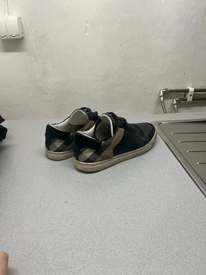 Svarta sneakers med rutig detalj - Säljer ett par svarta sneakers från Burberry med klassisk rutig panel i beige och svart på hälen. Skorna har snörning, rund tå och platt sula. Materialet är en mix av skinn och textil, med diskreta röda detaljer vid snörningen. Perfekt för dig som gillar stilrena och ikoniska designer. Skorna har defekter och ingen kartong eller påse fås med därav det billiga priset 