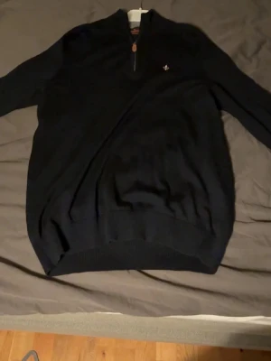 Svart merinoullströja från Morris - Säljer en svart långärmad tröja från Morris i 100% merinoull. Tröjan har en half zip-dragkedja med läderdetalj och en liten broderad logga på bröstet. Perfekt för dig som gillar stilrena och klassiska plagg.