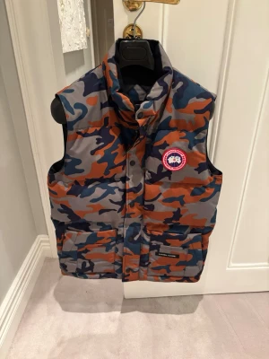 Camouflage dunväst från Canada Goose - Säljer en jättesnygg dunväst från Canada Goose i camomönster med blå, orange och grå toner. Västen har hög krage, två stora fickor framtill och klassisk Canada Goose-logga på bröstet. Perfekt för dig som vill sticka ut med streetstyle-vibe.