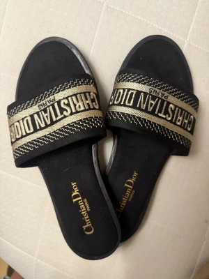 Svarta sandaler - Snygga svarta sandaler med bred rem i svart och guld där loggan är broderad. Platt sula och öppen tå, perfekt för sommaren. Tillverkade i textil och syntetmaterial med exklusiv känsla, liknande mockamatrial på insidan och fin design. Aldrig använt 💕