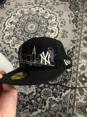 Svart New Era NY keps 59FIFTY - Säljer en svart New Era 59FIFTY keps med broderat New York Yankees-logo och vit stadssilhuett runt hela kepsen. Klassisk flat brim och detaljerad brodyr med bro och skyskrapor. Märkt Genuine Merchandise på insidan.