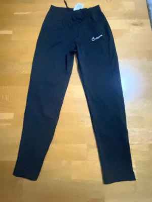Svarta Nike Dri-Fit träningsbyxor XL - Svarta träningsbyxor från Nike med Dri-Fit-teknologi och broderad vit Swoosh-logga på benet. Byxorna har elastisk midja med snörning och raka ben. Perfekta för sport eller chill. Tillverkade i lätt syntetmaterial.