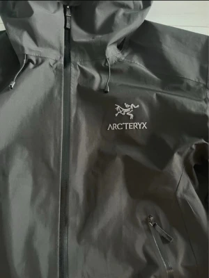 Svart Arc'teryx vindjacka Gore-Tex - Arc'teryx vindjacka i svart med huva och dragkedja framtill. Tillverkad i slitstark Gore-Tex Pro som står emot regn och vind. Jackan har två bröstfickor med dragkedja och justerbara muddar. Perfekt för dig som vill ha en clean och funktionell look.
