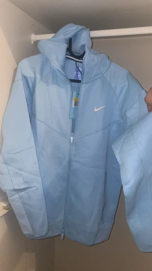 Nike NOCTA ljusblå hoodiejacka S - Nike NOCTA hoodiejacka i ljusblått med dragkedja, huva och vita loggor på bröstet och ärmen. Jackan har en clean look med snygga sömdetaljer och är tillverkad i mjuk bomullsmix. Perfekt för dig som gillar streetwear och vill sticka ut med en fräsch färg.