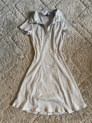 Ljusgrå ribbad kortklänning Divided - Supersnygg ljusgrå ribbad kortklänning från Divided. Klänningen har krage, korta ärmar och knappar vid halsen. Den är figurnära och gjord i mjuk bomull, perfekt för en chill och trendig look.