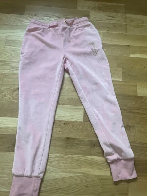 Rosa mjukisbyxor från Juicy Couture - Säljer ett par Rosa  mjukisbyxor från Juicy Couture i mjuk velour. Byxorna har resår i midjan, muddar vid bensluten och ett glittrigt JC-emblem med krona i guld på ena benet. Perfekta för chill dagar eller när du vill ha en extra cozy look.