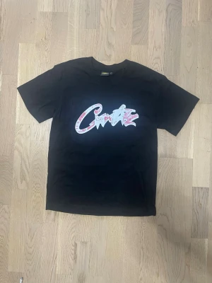 Svart Corteiz t-shirt med tryck - Snygg och trendig Corteiz t-shirt i klassisk svart färg med det ikoniska Corteiz-trycket på bröstet. Ett eftertraktat streetwear-plagg som sticker ut och passar perfekt till en modern vardagslook.  • Märke: Corteiz • Typ: T-shirt • Färg: Svart • Passform: Regular fit • Skick: Nytt skick • Coolt och unikt tryck framtill • Bekväm och mjuk kvalitet  Lätt att styla med jeans, cargos eller shorts. Perfekt för streetwear, vardag och sommarens outfits. Ett plagg med hög efterfrågan – först till kvarn!