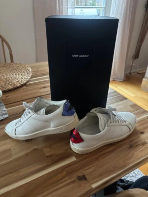 Saint Laurent vita sneakers med läppar - Säljer ett par vita Saint Laurent sneakers i skinn med coola detaljer i form av läppar på hälen, en i rött och en i blått. Väldigt bra skick. Storlek 40 men passar upp mot 42. Så inte true to size. 