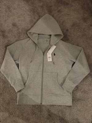 Grå hoodie från Polo Ralph Lauren - Snygg grå hoodie från Polo Ralph Lauren med dragkedja och huva. Tröjan har broderad svart logga på bröstet, känguruficka och ribbade muddar. Perfekt för chill dagar och enkel att matcha med jeans eller joggers.
