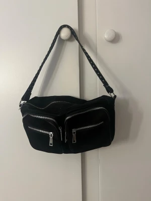 Svart axelväska från Noella i mocka - Säljer en svart axelväska från Noella i mjuk mocka. Väskan har två stora framfickor med silvriga dragkedjor och ett brett axelband med nitar. Perfekt för dig som vill ha plats för allt viktigt och ändå se cool ut. (Pris kan eventuellt diskuteras)