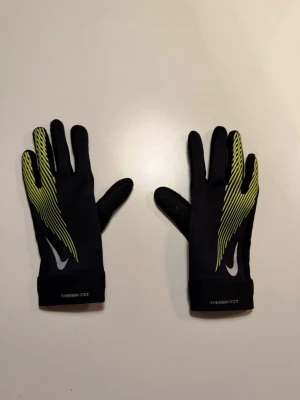 Svarta Nike Therma-FIT löparhandskar - Säljer ett par svarta Nike Therma-FIT löparhandskar med gulgröna detaljer och vita loggor. Handskarna har greppmönster i handflatan och är tillverkade i ett stretchigt syntetmaterial som håller värmen. Perfekta för löpning eller träning utomhus. Säljer för att dem är för små.