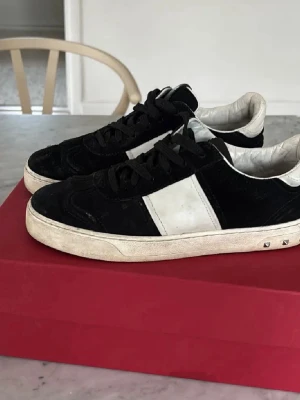 Valentino Garavani svarta sneakers - Säljer ett par svarta och vita Valentino flycrew sneakers i skinn och mocka. Skorna har klassisk snörning, rund tå och coola nitar på sulan baktill. Perfekta för dig som vill ha en stilren men edgy look.