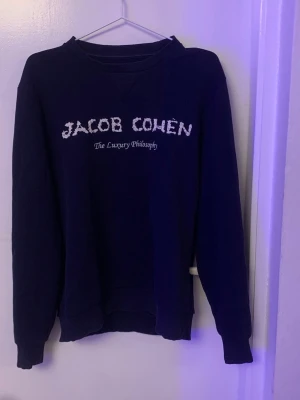Mörkblå sweatshirt från Jacob Cohën - Säljer en mörkblå sweatshirt från Jacob Cohën med vit broderad logga och texten 'The Luxury Philosophy' på bröstet. Tröjan har rund hals, långa ärmar och ribbade muddar. Perfekt för dig som gillar stilrena och exklusiva plagg.