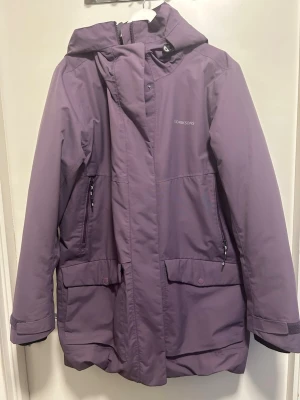 Lila parkas från Didriksons, strl 48 - Säljer en lila parkas från Didriksons i storlek 48. Jackan är 100% vattentät med huva, dragkedja och stora fickor framtill. Materialet är polyamid och polyester, perfekt för kalla och regniga dagar. Snygg och funktionell design med justerbara ärmslut.
