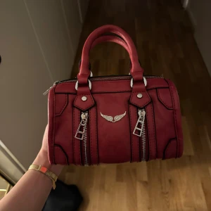 Zadig liknande väska - Zadig liknande handväska i skinnimitation med två rejäla handtag, dragkedjor framtill och en snygga silverfärgade detaljer. Den har ett rymligt fack med innerfickor och beige foder. Perfekt storlek för det viktigaste och ger en edgy vibe till din outfit. Använd ca 2 gånger