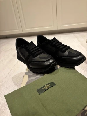 Valentino Rockrunner svarta sneakers - Svarta Valentino Rockrunner sneakers med coolt kamouflagemönster och detaljer i både skinn och mocka. Snygg platt sula med nitar på hälen och klassisk snörning. Perfekt för dig som gillar exklusiva och stilrena sneakers med edge.  Använd ett fåtal gånger men är för små för mig