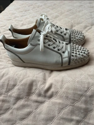 Vita sneakers med nitar från Loubouitin - Säljer ett par vita sneakers från Christian Louboutin med ikonisk röd sula och silverfärgade nitar över tån. Skorna har snörning, rund tå och är tillverkade i skinn med beige innersula. Perfekta för dig som vill sticka ut med en edgy detalj.