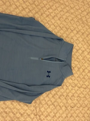 Blå half zip tröja från Under Armour - Säljer en ljusblå långärmad tröja från Under Armour med half zip och broderad logga på bröstet. Tröjan har loose fit och är gjord i ett mjukt syntetmaterial som passar perfekt till träning eller chill. Hög krage och ribbade detaljer vid halsen.