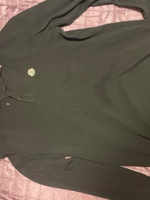 Svart långärmad polotopp Moncler - Säljer en svart långärmad polotopp från Moncler med klassisk krage och knappar framtill. Toppen har Monclers ikoniska logga på bröstet och är tillverkad i mjuk bomull som känns skön mot huden. Perfekt för dig som gillar stilrena och tidlösa plagg. NY PRIS LIGGER PÅ RUNT 4000KR