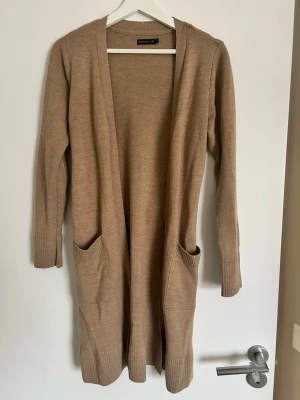 Beige kofta från Stockh LM med knytband - Lång beige kofta från Stockh LM med ribbade muddar, stora fickor framtill och ett snyggt knytband i midjan. Perfekt för lager på lager och chill stil. Materialet är mjukt och stickat, vilket gör den extra skön att bära.