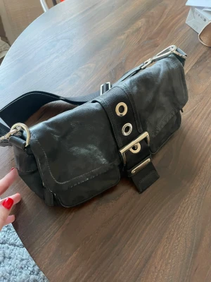 Svart axelväska från Prada i nylon - Säljer en svart axelväska från Prada med ikonisk metallogo på sidan. Väskan är gjord i nylon med detaljer i skinn och har en bred axelrem. Framsidan pryds av en stor metallspänne och dekorativa öljetter. Perfekt för dig som gillar en edgy och stilren look.