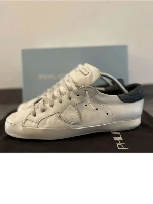 Vita sneakers från Philippe Model - Snygga vita sneakers från Philippe Model med svart detalj på hälen och klassisk snörning. Skorna har en rund tå, diskret logga på sidan och är tillverkade i skinn. Perfekta för dig som gillar stilrena och tidlösa sneakers.