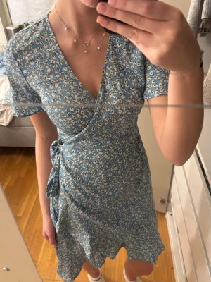 Blommig blå omlottklänning - Supersöt blå omlottklänning med vitt blommönster och knyt i midjan. Klänningen har korta ärmar och v-ringad hals. Från Only i storlek 36