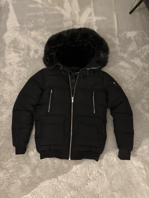 Svart pufferjacka Supply & Demand M - Säljer en svart pufferjacka från Supply & Demand med stor huva och fejkpäls runt kanten. Jackan har två stora fickor med lock, två dragkedjefickor framtill och ribbade muddar. Snygg patch på ärmen och hel dragkedja framtill. Perfekt för kalla dagar!