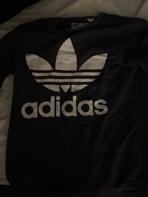 Svart sweatshirt från Adidas - Säljer en svart sweatshirt från Adidas med det klassiska vita Trefoil-loggan tryckt stort på bröstet. Tröjan har rund halsringning och långa ärmar. Tillverkad i mjuk bomull för skön känsla och stilren look.