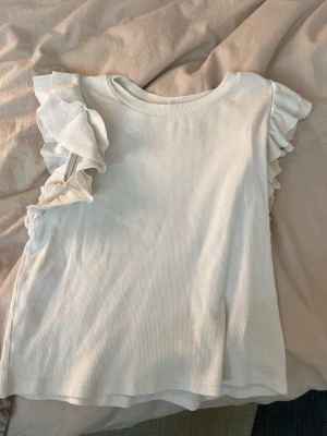 Vit ribbad topp med volangärmar - Säljer en söt vit topp med ribbad struktur och volangdetaljer på ärmarna. Toppen är ärmlös med breda axelvolanger som ger en extra touch. Perfekt till jeans eller kjol för en fräsch look. Från H&M