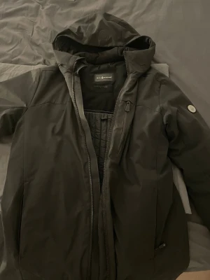 Svart vinterjacka från Sail Racing - Säljer en svart vinterjacka från Sail Racing i storlek Large. Jackan har huva, dragkedja framtill, bröstficka med dragkedja och diskret logga på ärmen. Materialet är slitstarkt och vattenavvisande, perfekt för kalla dagar. Snygg och funktionell design som håller dig varm.