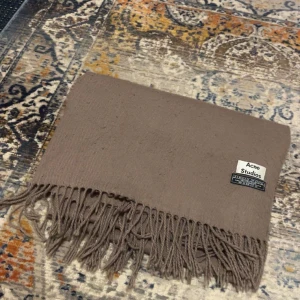 Beige halsduk från Acne Studios - Säljer en stilren beige halsduk från Acne Studios. Halsduken är i mjuk ull och har klassiska fransar i kanterna samt en diskret logotyp. Perfekt accessoar för att lyfta din outfit under kyliga dagar. Fint skick.