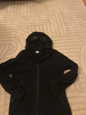 C.p company cardigan  - Säljer en stilren C.P. Company hoodie i svart med den ikoniska goggle-huvan. Tjock och bekväm kvalitet som passar perfekt som lätt hoodie under lite kyligare dagar dagar.