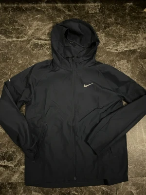 Nike Miller  - Säljer en svart vindjacka från Nike med huva och dragkedja framtill. Jackan har en stilren look med Nike-logga på bröstet och praktiska fickor på sidorna. Perfekt för blåsiga dagar och enkel att matcha med olika outfits.