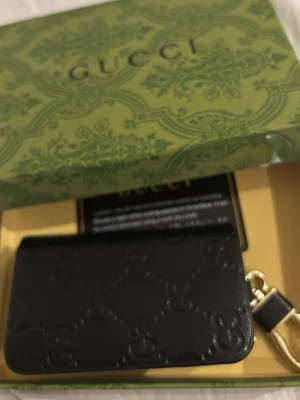 Svart  plånbok i skinn - Säljer en svart plånbok från i äkta skinn med mönster i relief. Plånboken har guldfärgade metalldetaljer och en kedja med ring för nycklar. Kommer i originalförpackning och är riktigt snygg att ha i väskan eller hänga på nyckelknippan.